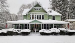 large green house with covered front porch, trees in the background, and bushes along the front. Everything is covered in white fluffy snow.