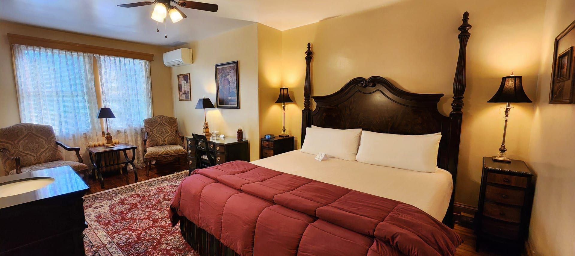 Guest room with king bed, wood dresser, sink with mirror and upholstered chairs by a large window