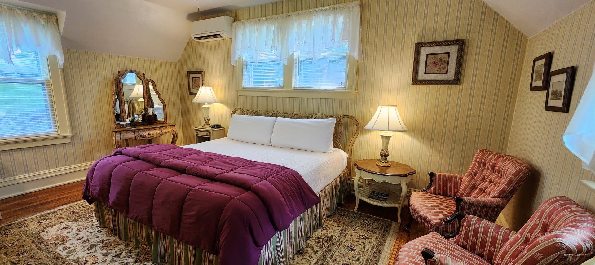 Pretty guest room with yellow striped wallpaper, king bed, dresser with mirror and striped sitting chairs. Large area rug over hardwood floors.