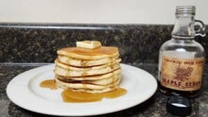 plate with a stack of pancakes with pat of butter on top drizzled with maple syrup. Bottle of maple syrup on right side of plate.